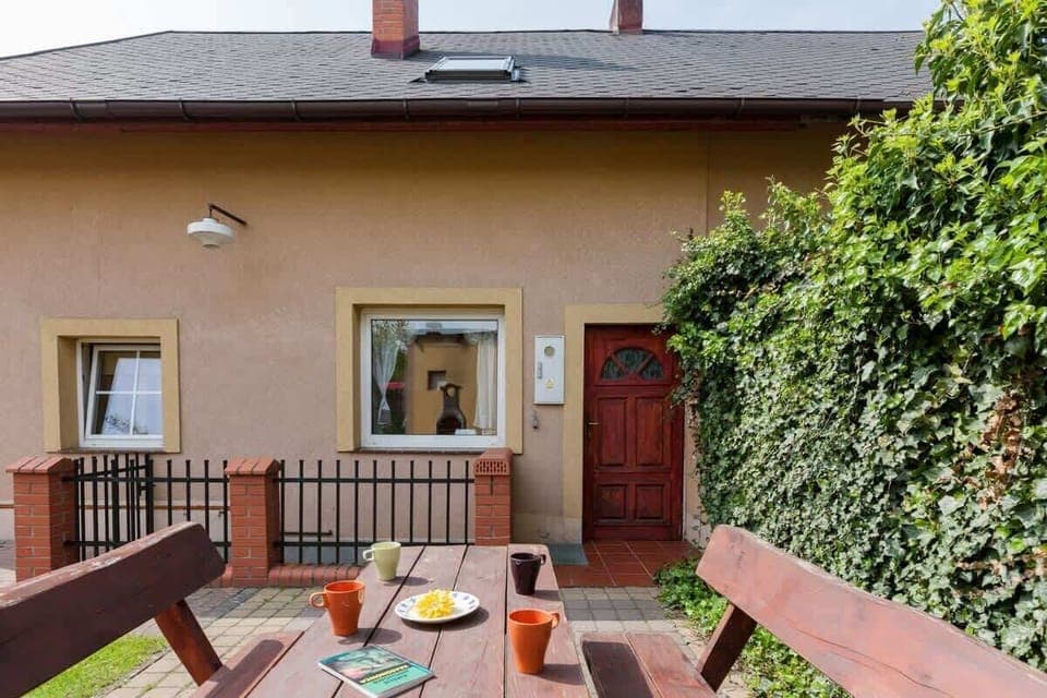 View of the house from the front. A wooden table with benches is visible in the foreground and the entrance to the house can be seen behind them. 