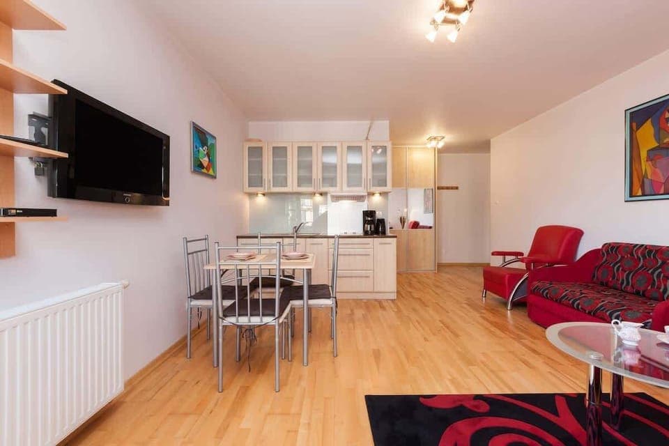 An open-plan room with a dining and living area. There is a dining table with four chairs and a red sofa and armchair. The kitchen is visible in the background with light wood cabinets.