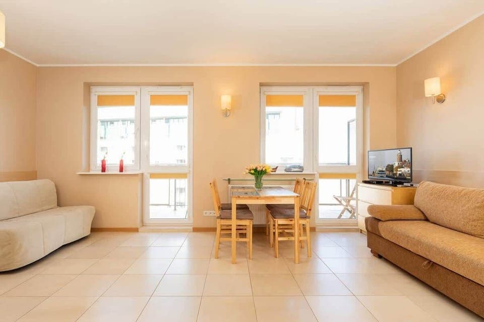 A bright living room with a beige sofa, large windows, and a small dining table. The space feels open and welcoming.