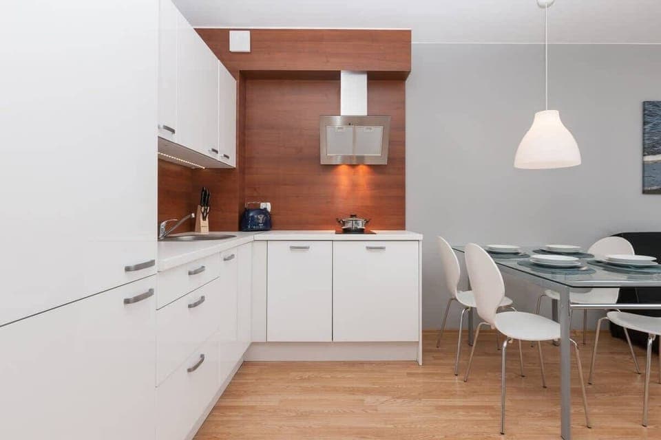 A clean, spacious kitchen with white cabinetry, a dining area with a wooden table and modern chairs. The kitchen features stainless steel appliances and hanging lights.