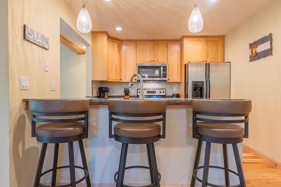 Kitchen with wooden cabinets, stainless steel appliances, and a breakfast bar with three brown padded bar stools. Pendant lights hang above the counter.