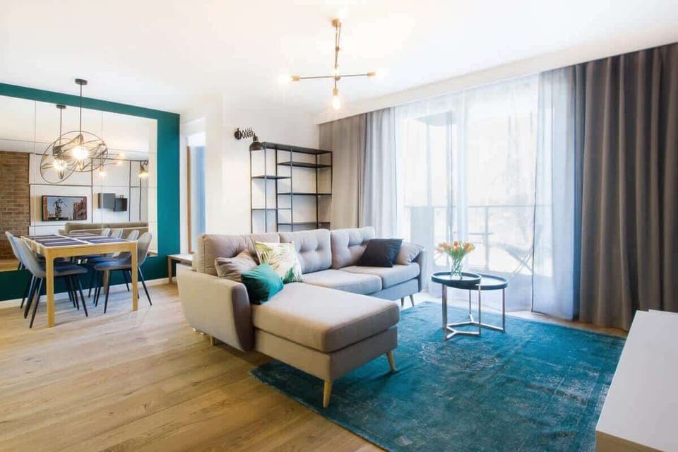 A stylish living room with a modern aesthetic, featuring a cozy beige sofa, vibrant teal rug and coffee table. The room is well-lit with large windows and decorated with tasteful wall art.