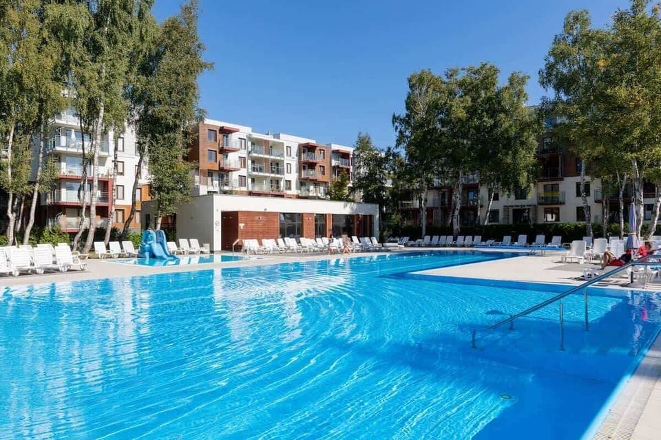A large outdoor pool with clear blue water, surrounded by greenery and apartment buildings in the background.