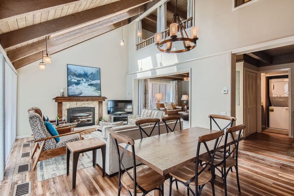 A cozy living and dining area with wooden flooring, a fireplace, a TV, and modern furnishings. A dining table with chairs is in the foreground, and a small laundry room is visible in the background.