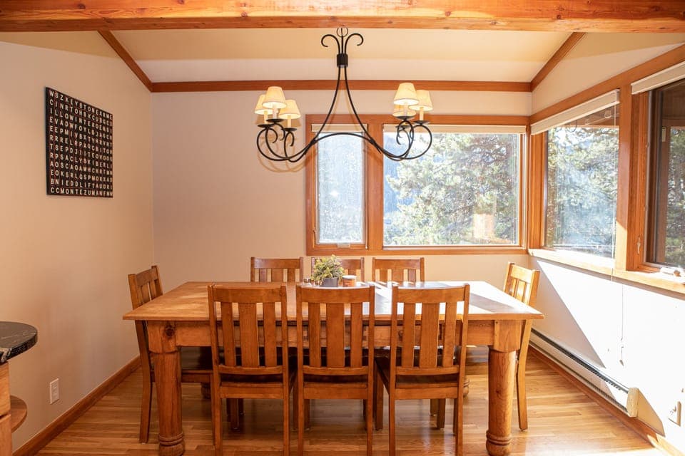 A wooden dining table with six matching chairs is under a chandelier in a room with large windows and natural light. A framed word search puzzle hangs on the wall.
