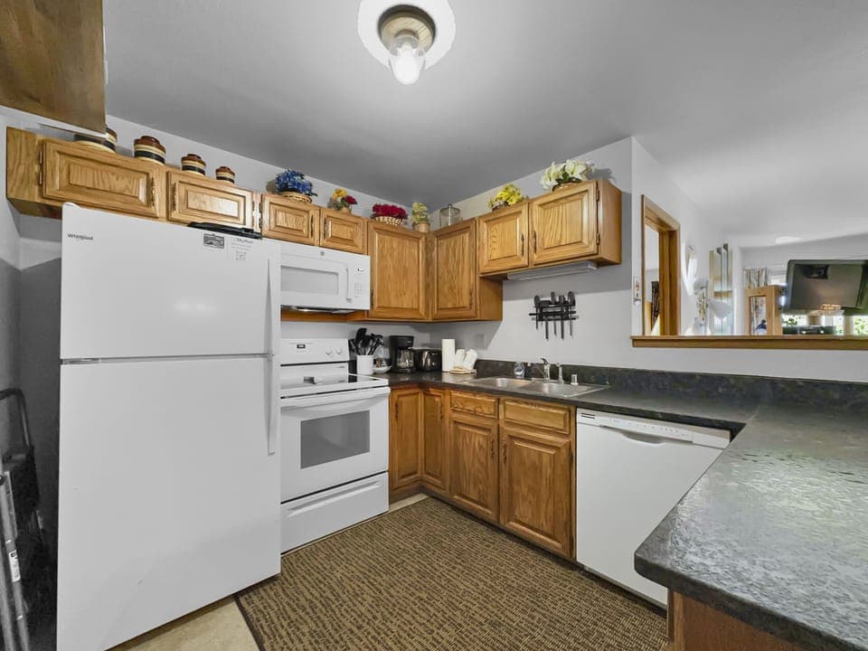 A kitchen with wooden cabinets, a white refrigerator, oven, and microwave, a dishwasher, and a dark countertop. A rug is placed on the floor, and various decorative items are on the upper cabinets.