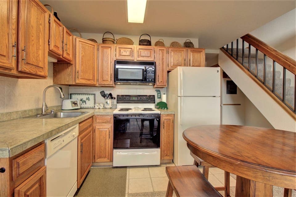 A small kitchen with wooden cabinets, a microwave, stove, and refrigerator. There is a sink, dishwasher, and a round wooden table with chairs. A staircase is visible in the background.