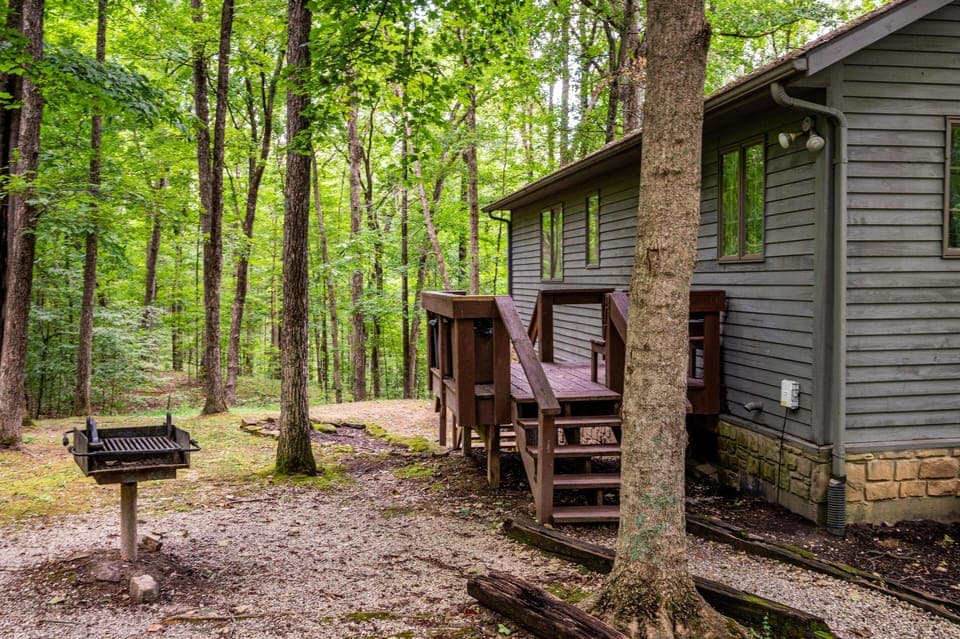 Side deck with dark wood railing and stairs leads to a wooded yard featuring a metal charcoal grill on a gravel patch.