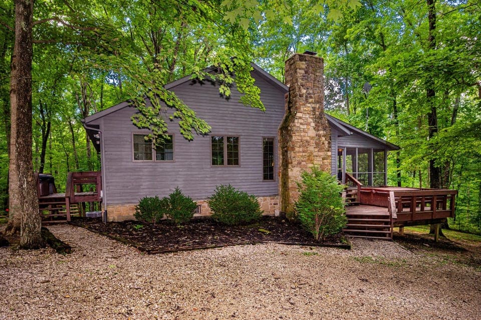 Gray wood-sided cabin exterior with stone chimney, screened porch, and raised deck surrounded by green trees.