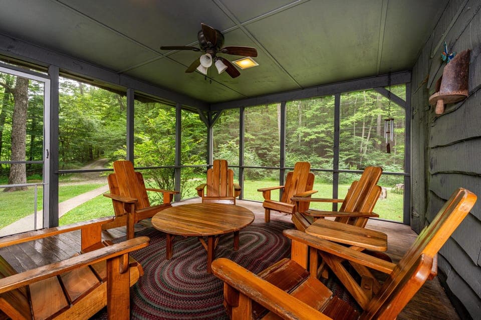 Screened living room porch with honey-toned Adirondack chairs, oval wood table, braided rug, and forest views.