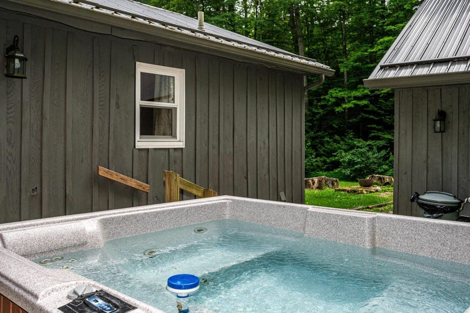 Outdoor hot tub with bubbling water sits between two gray wood-paneled buildings, overlooking a grassy yard and firepit.