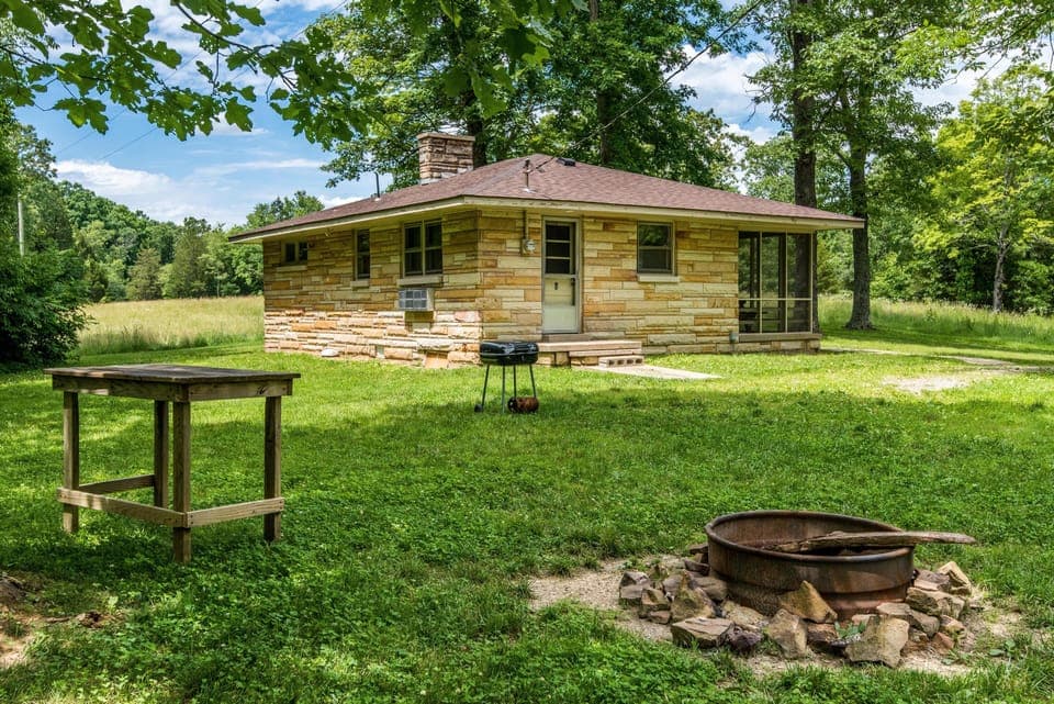 Outdoor fire pit encircled by stones with a charcoal grill and wooden prep table on green lawn.