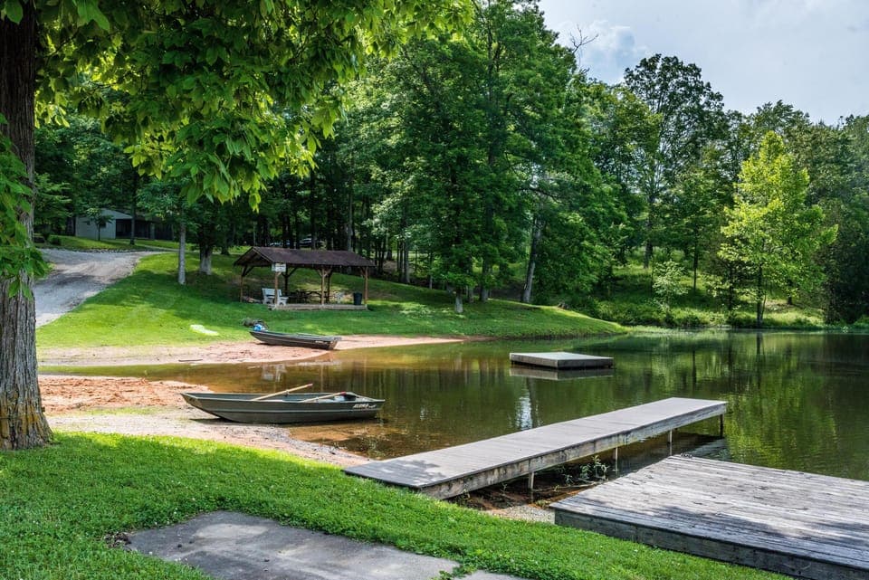Calm lake shore with two green rowboats, wooden docks, sandy beach, and shaded picnic pavilion nearby.