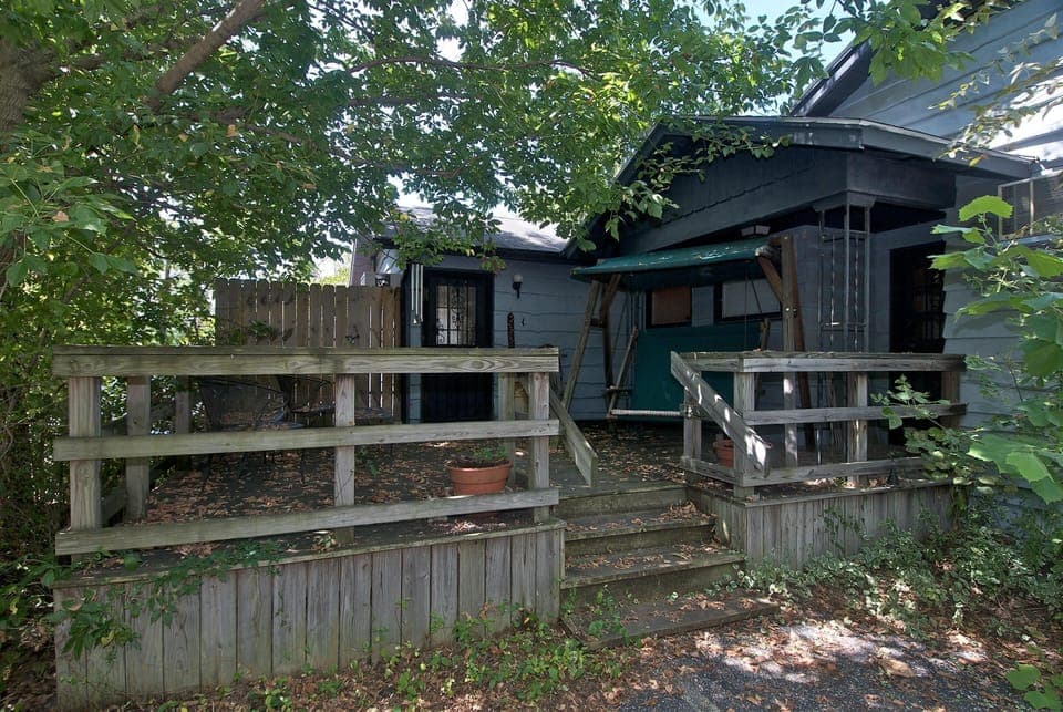 Shaded wooden deck with railing, green canopy porch swing, and black metal patio chairs surrounded by trees.