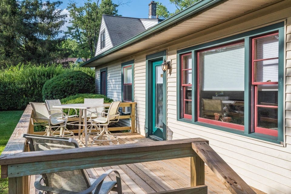 Sunlight illuminates the wooden deck with green-trimmed windows and red window frames beside a teal door.