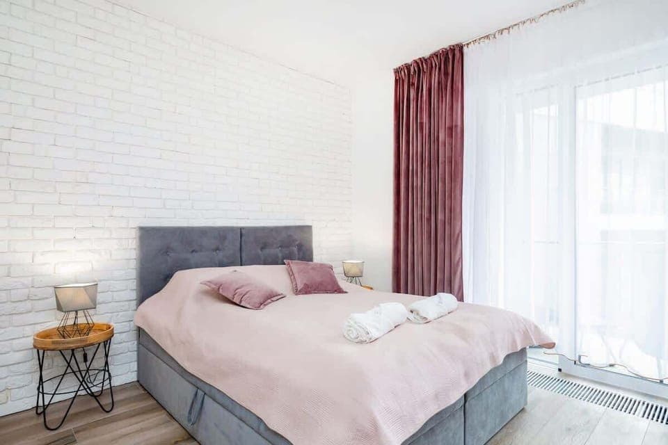 A bright and cozy studio featuring a large bed with light gray and pink bedding. A soft area rug and bedside tables with modern lamps complement the decor, with natural light streaming through a window with sheer curtains.