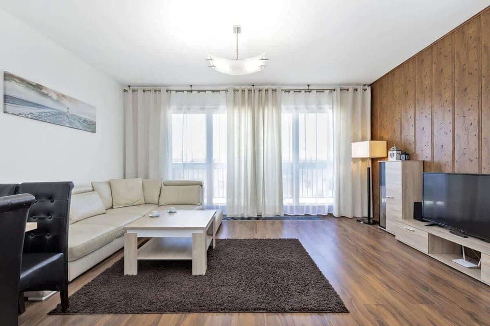 A spacious living room with a sofa, coffee table, TV, and a standing lamp. The interior is bright, with a large window and curtains, and there is a rug on the floor.