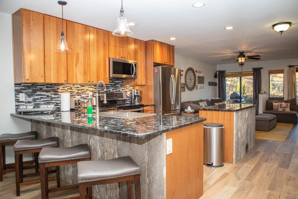 Modern kitchen and living area with wooden cabinets, granite countertops, stainless steel appliances, an island with barstools, a ceiling fan, and a wall-mounted dartboard.