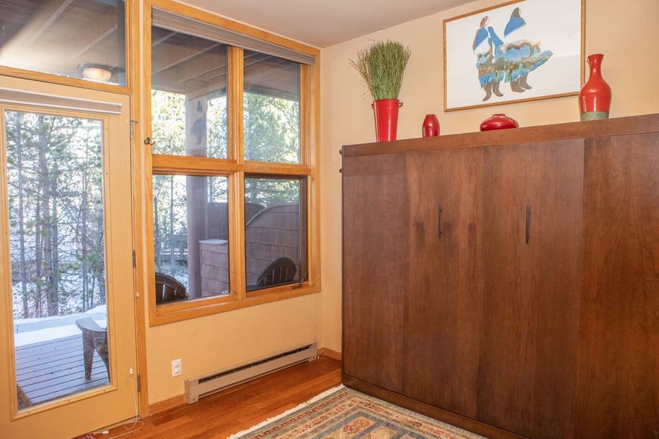 A room with a large wooden cabinet, a window with forest view, colorful rug, red vases on the cabinet, and a framed artwork on the wall.