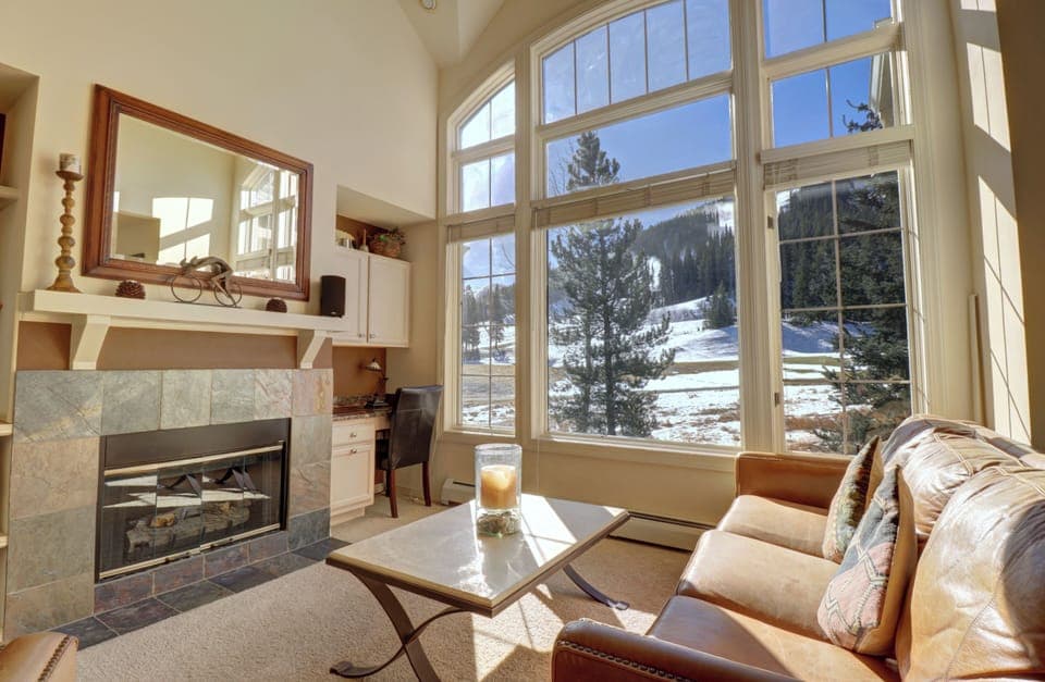 Spacious living room with large windows offering a view of the snowy East Village ski slopes. Features a fireplace, a cozy sofa, and coffee table. Natural light fills the room.