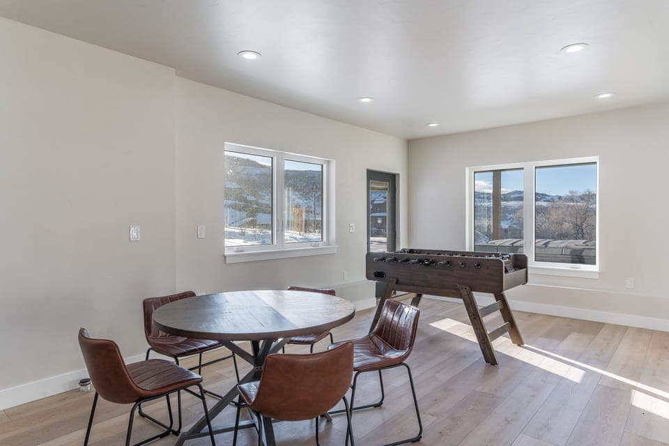 A room with a round wooden table and five brown chairs on a light wood floor. There is a foosball table near windows showing an outdoor view.