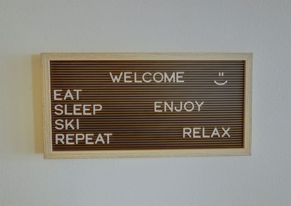 A wooden-framed sign with white letters reads, "WELCOME. EAT SLEEP SKI REPEAT ENJOY RELAX" on a brown background. There is a smiley face next to the word "WELCOME.