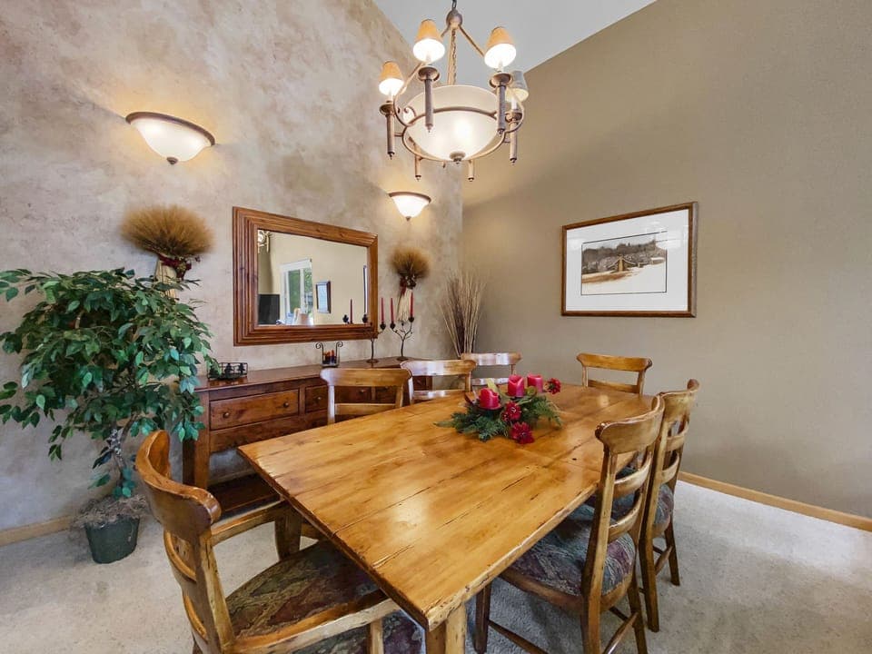 Dining room with a wooden table set for six, chandelier, framed artworks on beige textured walls, potted plant, sideboard with candles, and minimal festive decor on the table.