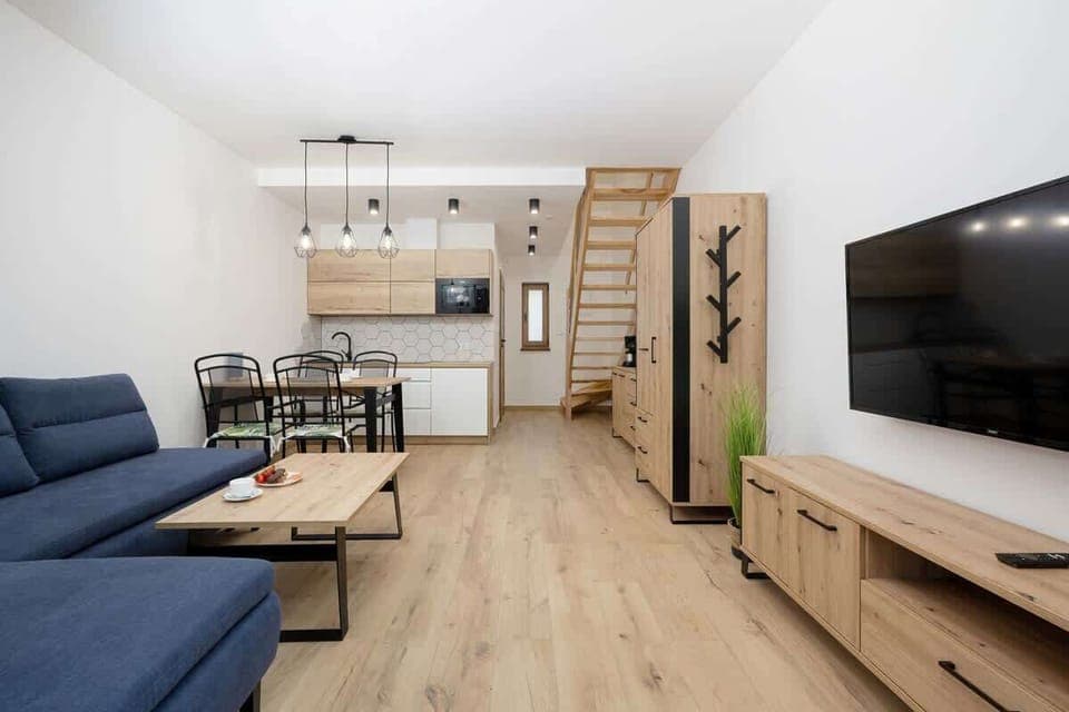 Another angle of the living room showing the dining area in the background. The layout is open-plan, with a small dining table and chairs near the back, maintaining a natural, wooden aesthetic throughout.