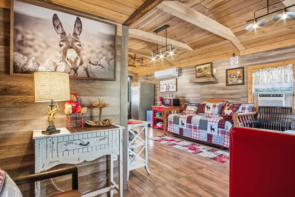 A cozy interior view of the cabin's living area, featuring a daybed with plaid bedding, a distressed white side table with an antler lamp, and a large, whimsical portrait of a donkey on the wood-paneled wall.