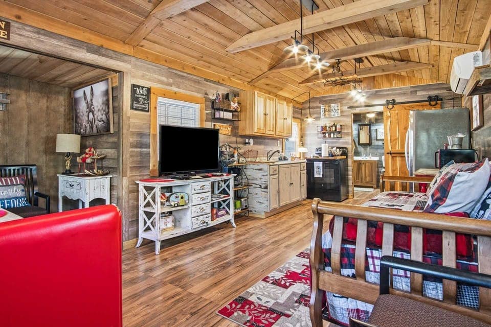 The interior of a cozy, open-concept cabin featuring vaulted wood-plank ceilings, a well-equipped kitchenette, and a living area with a television, red accents, and rustic wooden finishes throughout.