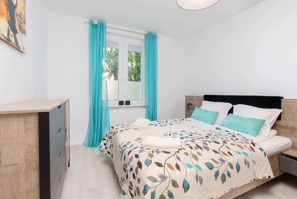 A cozy bedroom with a patterned bedspread, turquoise curtains, and bright natural lighting.
