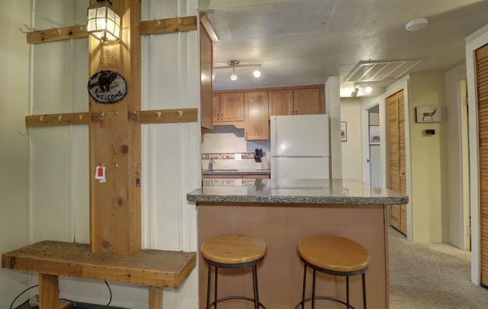 A kitchen with wooden cabinets, a white refrigerator, granite countertops, and two wooden barstools. A hallway and closet doors are visible. There's a welcome sign and coat hooks on the left wall.