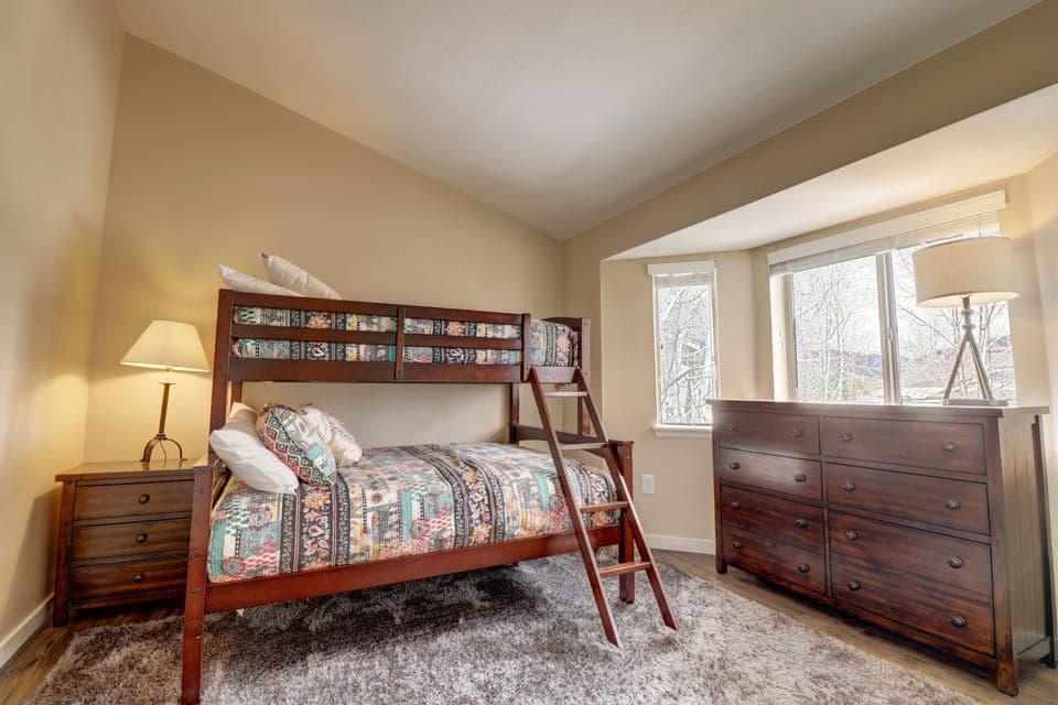 A bedroom with a wooden bunk bed featuring a colorful patterned bedspread, a wooden dresser, a nightstand with a lamp, and a window with shades partially open.