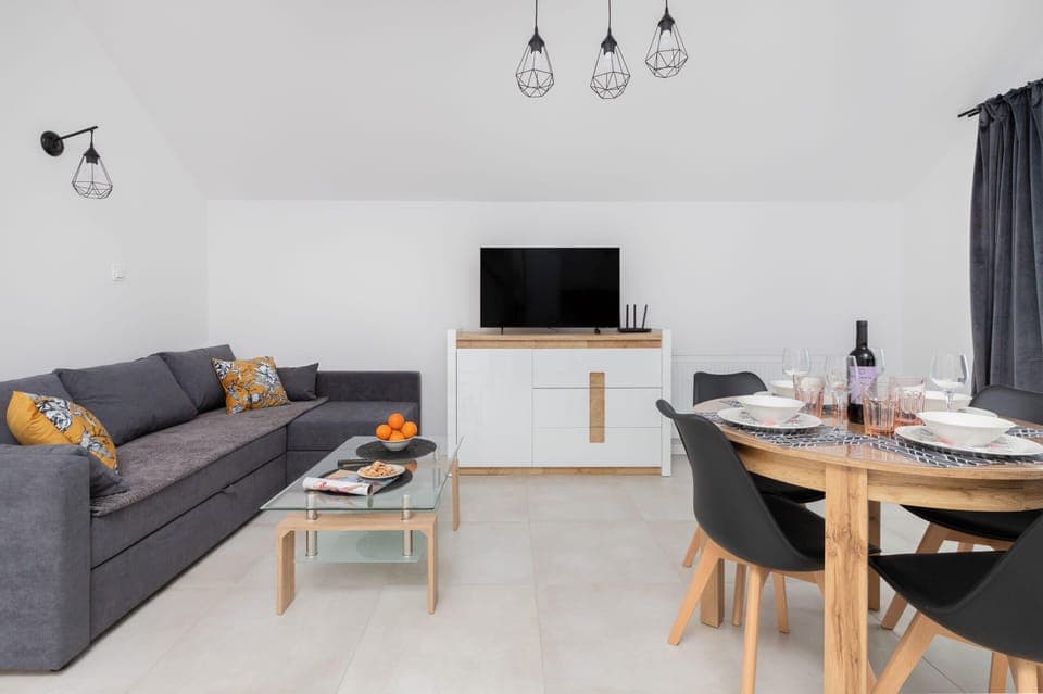A cozy living room with a gray sofa, small coffee table, and wall-mounted TV. The space is complemented by large windows with sheer curtains.