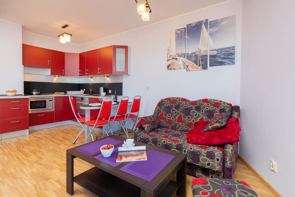 Open-plan living and dining area with red kitchen accents, wooden floors, and modern lighting.
