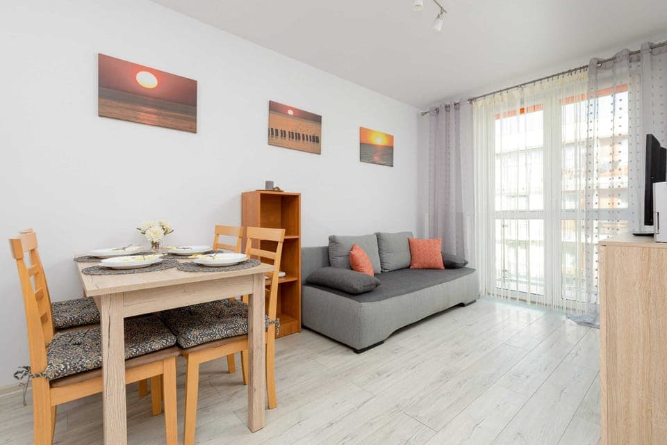 A living room with bright walls, a large sofa, a coffee table, and paintings on the wall. A spacious window allows a lot of natural light into the room.