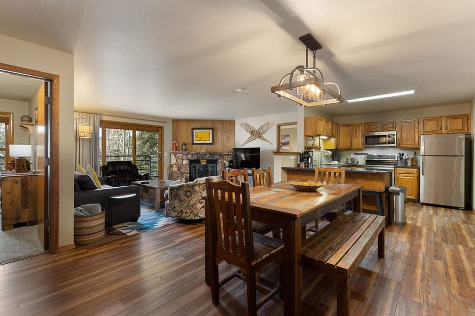 A cozy open-plan living area with a dining table, kitchen, and lounge featuring a fireplace and TV. Wood furnishings and warm lighting create a rustic feel. Large windows offer natural light.