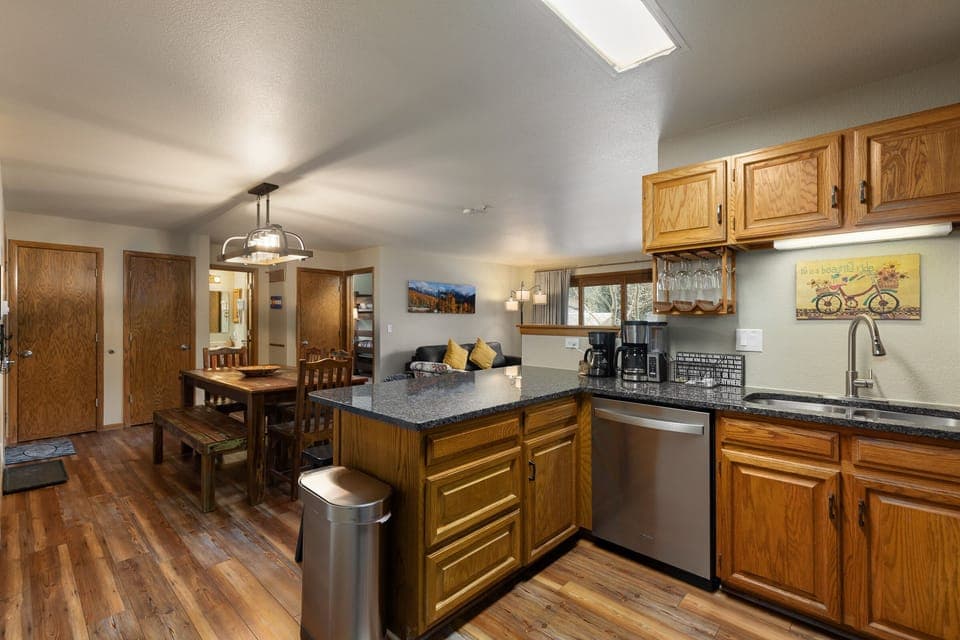 A modern kitchen with wooden cabinets, a granite countertop, stainless steel appliances, and a dining area in the background featuring a wooden table and chairs on wood flooring.