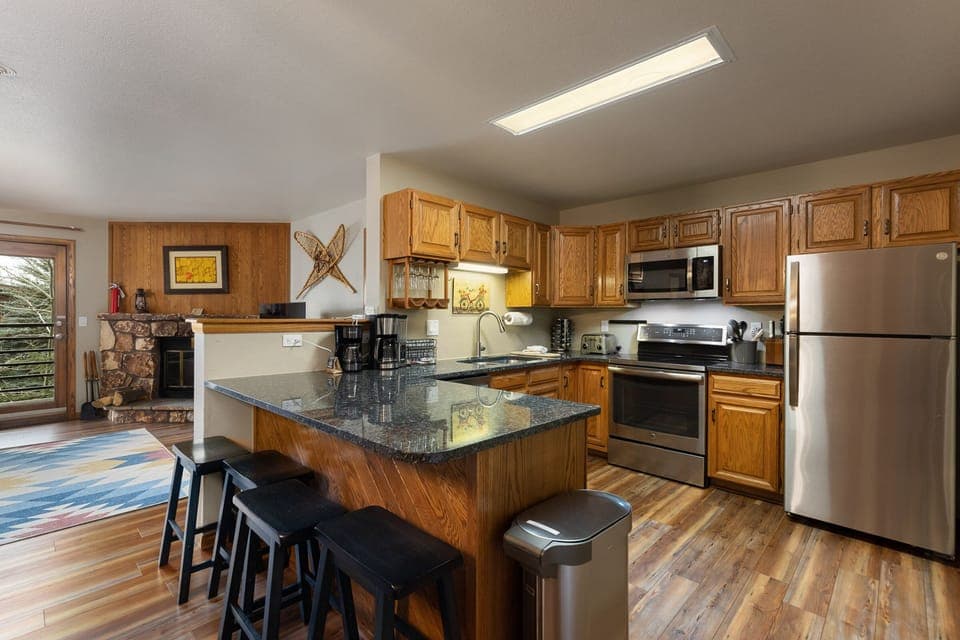 A kitchen with wooden cabinets, stainless steel appliances, a granite countertop island with three black stools, and a hardwood floor. A living area with a fireplace is visible in the background.