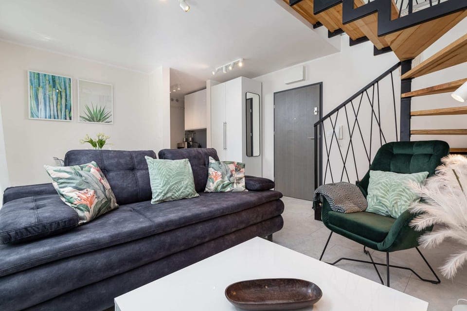 A cozy living area featuring a grey sofa, green armchair, and wooden staircase. The space is decorated with modern furniture and accents.