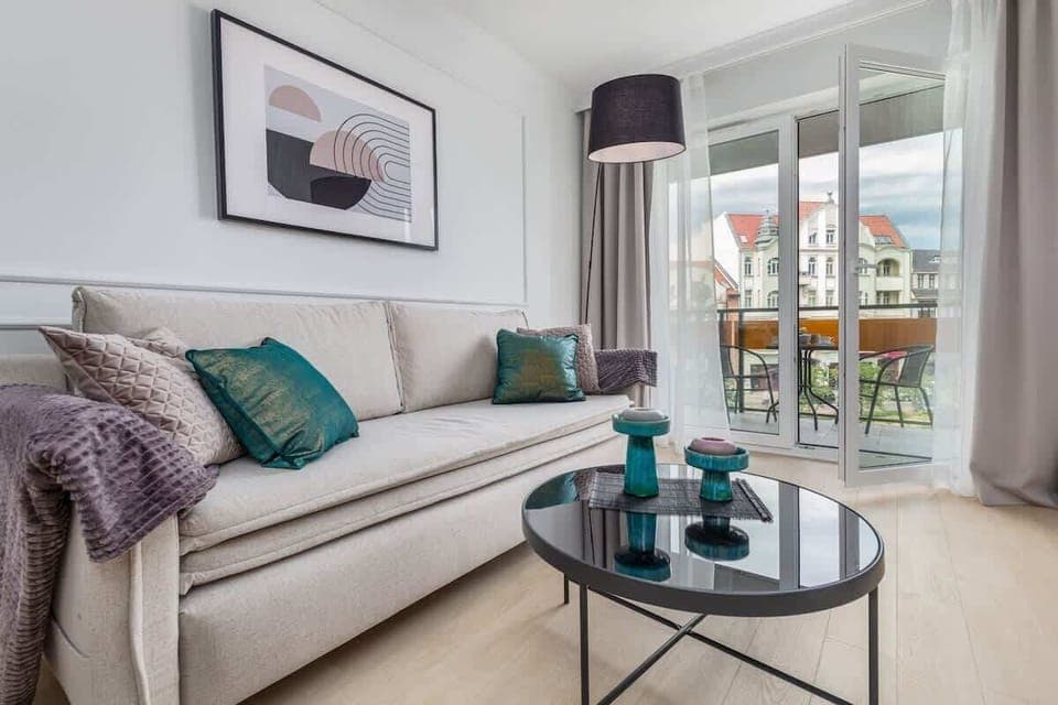 A stylish living room with a beige sofa, decorative pillows, a round coffee table, and a floor lamp, complemented by a balcony with a view of the city.

