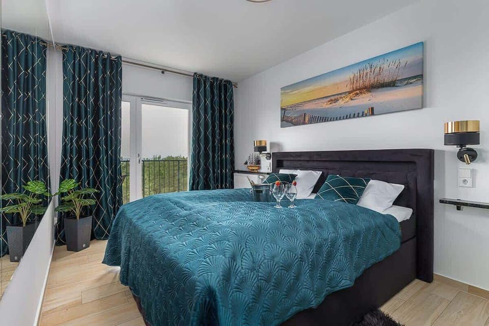 A double bed with deep green bedding, bedside tables with lamps, and a large window with thick drapes.