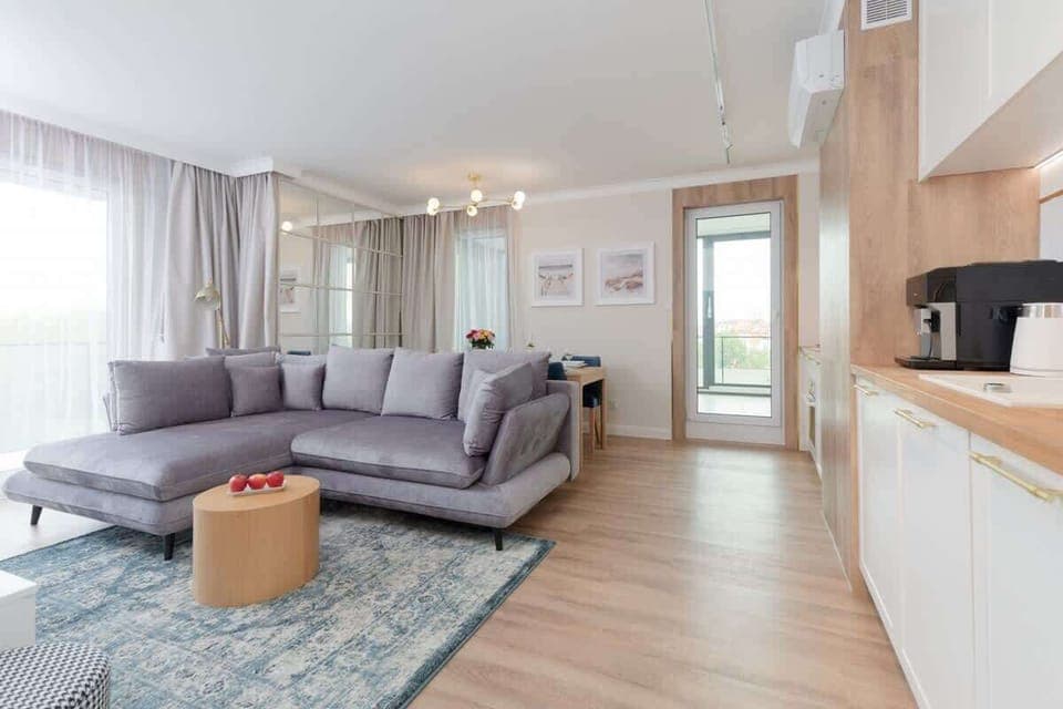 A spacious living room with a gray sofa, light wood floors, and a coffee table. The room is brightly lit by large windows, offering a cozy yet modern ambiance.
