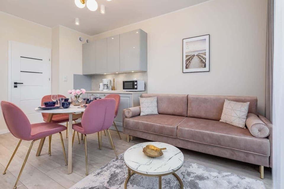 A bright and modern living room with a pink sofa, a marble coffee table, and a compact kitchen in the background.
