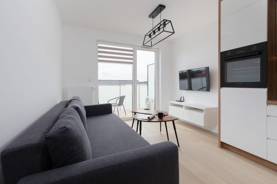 A modern living room featuring a gray sofa, wooden coffee table, and a wall-mounted TV, with bright natural lighting from large windows.