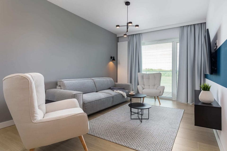 A stylish living room featuring a comfortable grey sofa, a coffee table, and large windows with curtains, creating a bright and inviting atmosphere.
