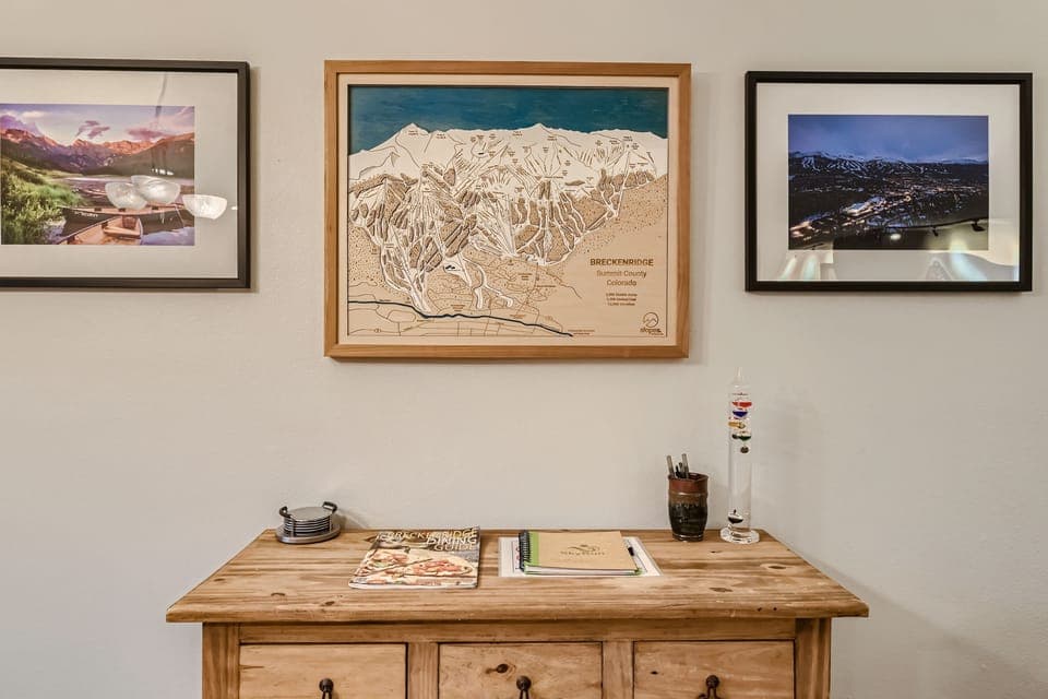 A wooden cabinet with a few magazines and small objects sits below a framed map of Breckenridge and two framed photographs of mountainous landscapes on a beige wall.