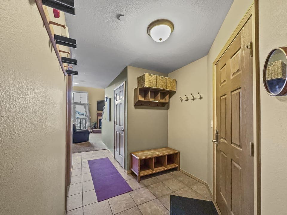 A hallway with tiled floors features a wall-mounted coat rack, a bench with storage, a purple rug, and a light fixture on the ceiling. The hallway leads to a living room in the distance.