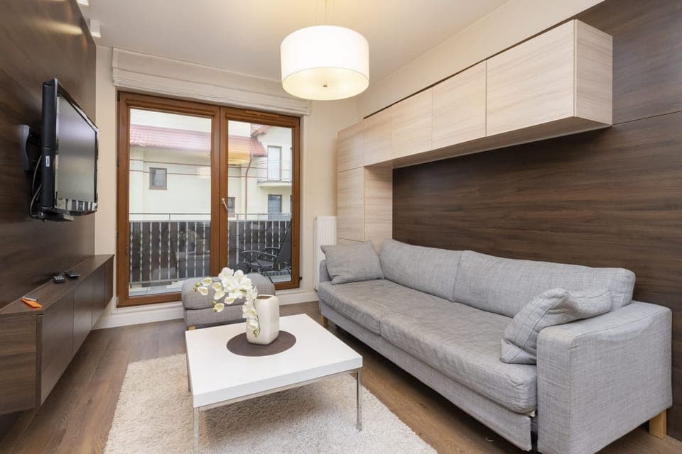 The living room features a modern gray sofa, a coffee table, and large windows that let in natural light. The space is cozy yet contemporary, with neutral tones and wooden flooring.