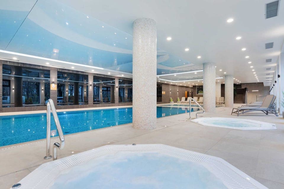 A luxurious indoor swimming pool area with a jacuzzi, surrounded by lounge chairs.
